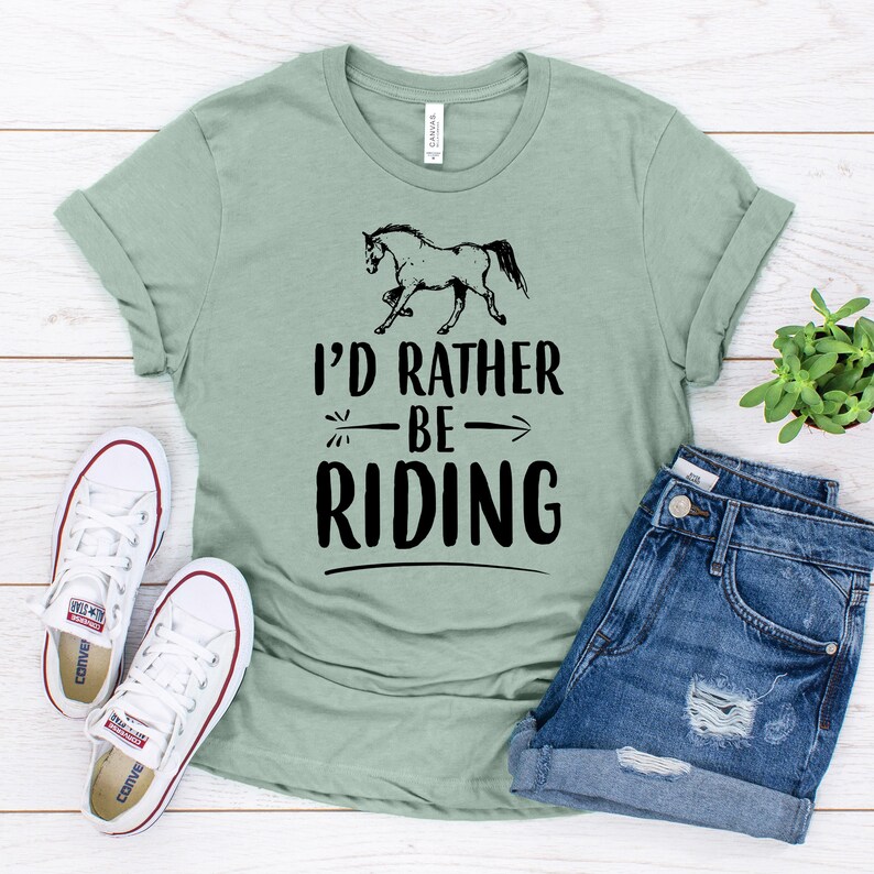 Puede incluir: Camiseta verde salvia con la frase "I'd Rather Be Riding" y un gr&aacute;fico de un caballo negro. La imagen incluye pantalones cortos de mezclilla y zapatillas de lona blancas, creando una est&eacute;tica ecuestre relajada.