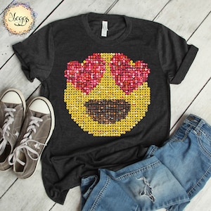 Puede incluir: Camiseta gris oscuro con un gran dise&ntilde;o de cara emoji. La cara emoji est&aacute; compuesta por emojis m&aacute;s peque&ntilde;os, con gafas de sol rosas en forma de coraz&oacute;n y una cara amarilla. Zapatillas grises y vaqueros azules est&aacute;n al lado de la camiseta.