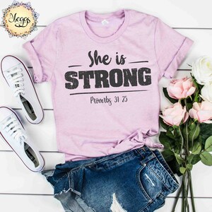 May include: A light purple t-shirt with the text "She is STRONG" in black with a distressed look. The text "Proverbs 31:25" is also printed in black below the main text.