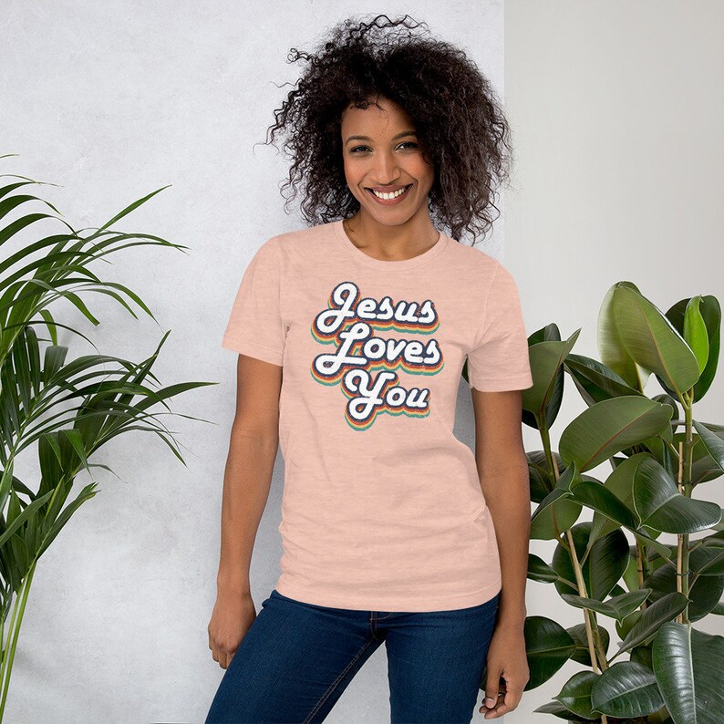 Puede incluir: Una camiseta de color rosa claro con un gr&aacute;fico de estilo retro que dice "Jesus Loves You" en colores arco&iacute;ris.