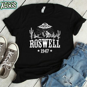 May include: Black t-shirt with a white graphic of a UFO over a desert landscape with cacti and mountains. The text "Roswell 1947" is below the graphic.