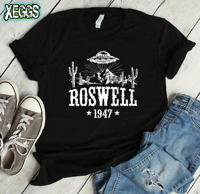 May include: Black t-shirt with a white graphic of a UFO over a desert landscape with cacti and mountains. The text "Roswell 1947" is below the graphic.