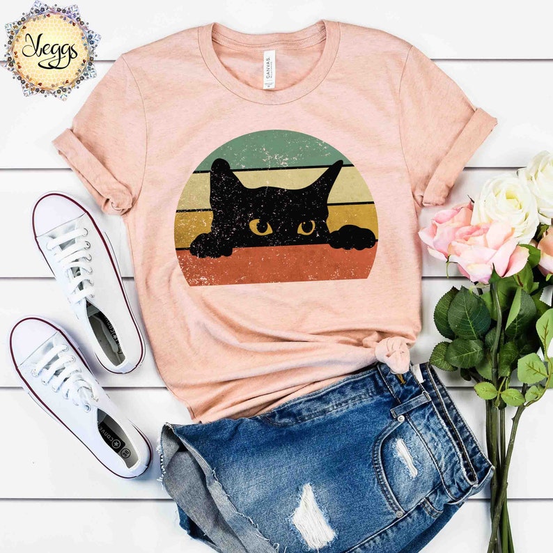 Puede incluir: Una camiseta de color melocot&oacute;n con un gr&aacute;fico de estilo vintage de un gato negro asom&aacute;ndose desde detr&aacute;s de una puesta de sol colorida.