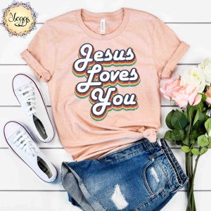 Puede incluir: Una camiseta de color melocot&oacute;n con un gr&aacute;fico de estilo retro que dice "Jesus Loves You" en colores arco&iacute;ris. La camiseta est&aacute; colocada sobre una superficie blanca con un par de zapatillas blancas y unos pantalones cortos de mezclilla azul.