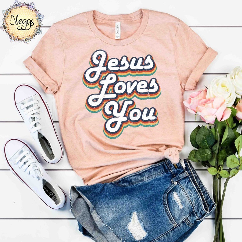 Puede incluir: Una camiseta de color melocot&oacute;n con un gr&aacute;fico de estilo retro que dice "Jesus Loves You" en colores arco&iacute;ris. La camiseta est&aacute; colocada sobre una superficie blanca con un par de zapatillas blancas y unos pantalones cortos de mezclilla azul.