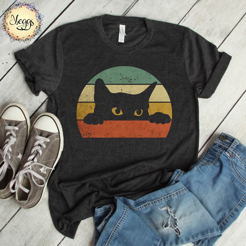 Puede incluir: Una camiseta gris oscuro con un gr&aacute;fico de estilo vintage de un gato negro asom&aacute;ndose sobre una puesta de sol colorida a rayas.