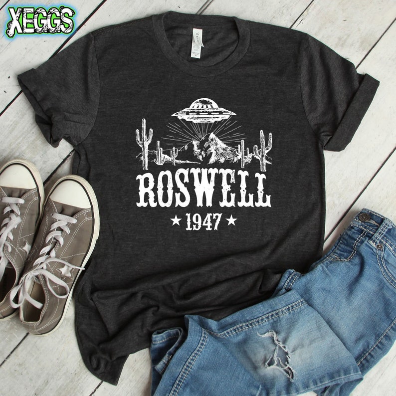 May include: A dark gray t-shirt with a white graphic of a UFO over a mountain range with cacti. The text "ROSWELL *1947*" is printed below the graphic.