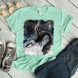 Puede incluir: Una camiseta verde claro con un estampado gr&aacute;fico de un hombre de las cavernas montando un monstruo marino en un mar tormentoso.