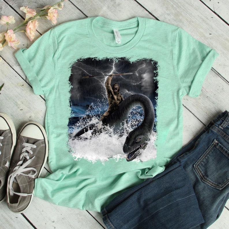 Puede incluir: Una camiseta verde claro con un estampado gr&aacute;fico de un hombre de las cavernas montando un monstruo marino en un mar tormentoso.