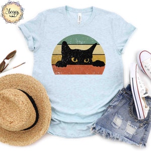 Puede incluir: Una camiseta azul claro con un gr&aacute;fico de estilo vintage de un gato negro asom&aacute;ndose desde detr&aacute;s de una puesta de sol colorida.
