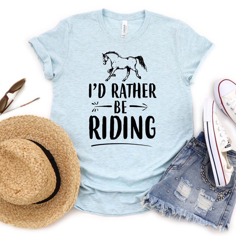 Puede incluir: Camiseta azul claro con el texto "I'd Rather Be Riding" y un dibujo de un caballo negro. La imagen incluye un sombrero de paja, gafas de sol, pantalones cortos vaqueros y zapatillas blancas.
