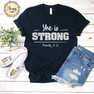 May include: Black t-shirt with white text that reads "She is STRONG Proverbs 31:25". The shirt is laid out on a wooden surface with a pair of white sneakers and denim shorts.