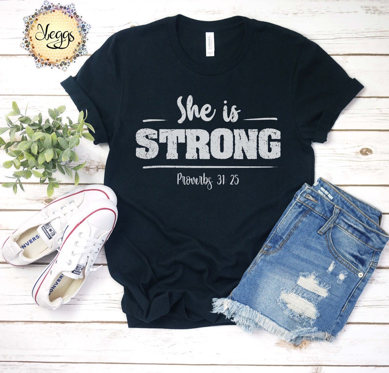 May include: Black t-shirt with white text that reads "She is STRONG Proverbs 31:25". The shirt is laid out on a wooden surface with a pair of white sneakers and denim shorts.