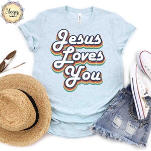 Puede incluir: Una camiseta azul claro con un gr&aacute;fico de estilo retro que dice "Jes&uacute;s te ama" en colores arco&iacute;ris.