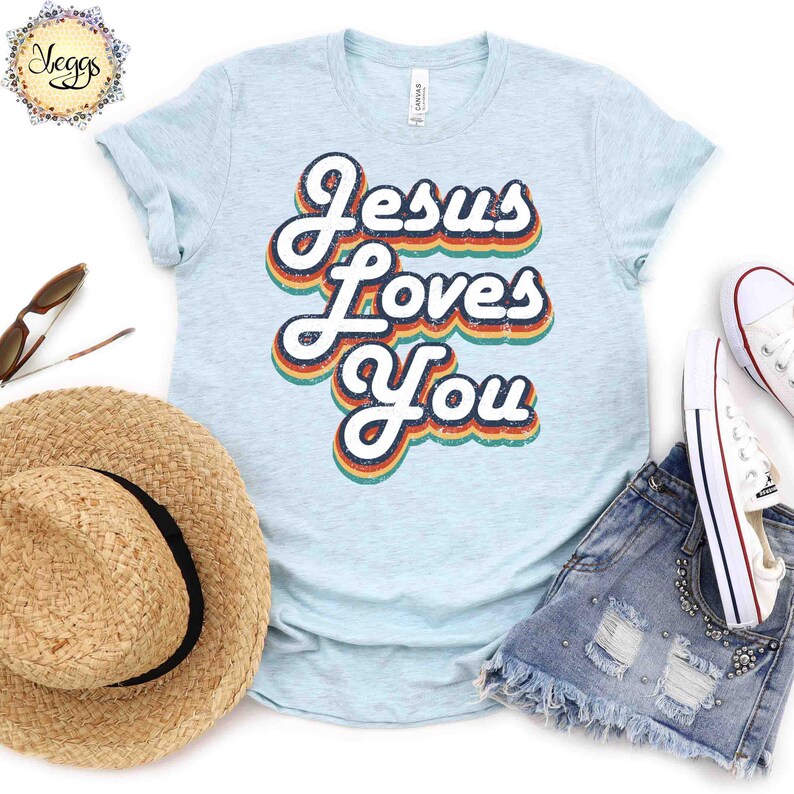 Puede incluir: Una camiseta azul claro con un gr&aacute;fico de estilo retro que dice "Jes&uacute;s te ama" en colores arco&iacute;ris.
