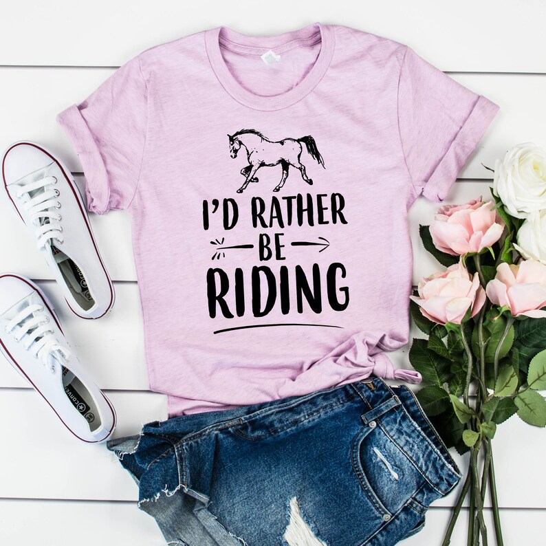 Puede incluir: Una camiseta de color lila claro con el texto "I'd Rather Be Riding" y un gr&aacute;fico de un caballo negro. La camiseta se combina con pantalones cortos de mezclilla y zapatillas blancas. Un ramo de rosas rosas tambi&eacute;n est&aacute; en la imagen.