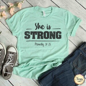 May include: A light green t-shirt with the text "She is STRONG" in black with a distressed look. Below the text is "Proverbs 31:25" in black.