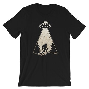 May include: Black t-shirt featuring a graphic of a UFO beaming down on a silhouette of Bigfoot in a forest. The UFO has two alien figures inside. The design is in a distressed, vintage style.