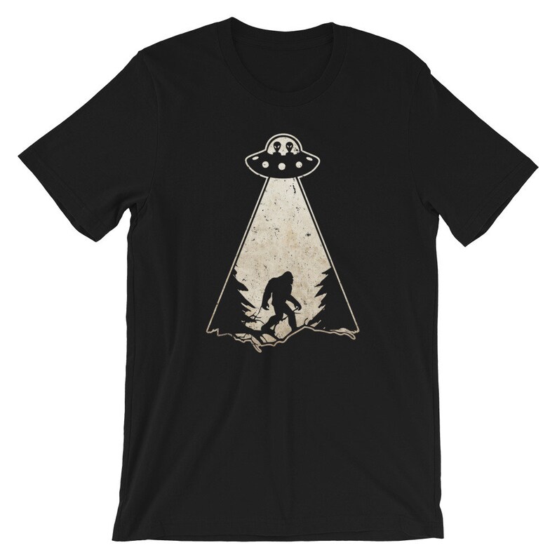 May include: Black t-shirt featuring a graphic of a UFO beaming down on a silhouette of Bigfoot in a forest. The UFO has two alien figures inside. The design is in a distressed, vintage style.