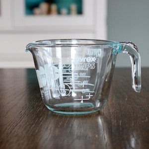 May include: Clear glass Pyrex measuring cup with a handle and measurement markings. The cup has a light blue rim and handle. The measurements are in cups and fractions of cups. The Pyrex logo is visible on the side.