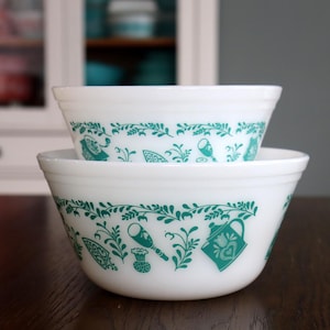 May include: Two white mixing bowls stacked. Each bowl features a teal green floral and kitchen utensil design around the rim. The bowls are on a dark brown surface, with a white cabinet in the background.