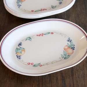 May include: Oval serving platters with a white background and a decorative fruit design. The border has a red and blue trim. The fruit design includes peaches, grapes, and berries. The platters are stacked on a dark wooden surface.