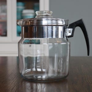 以下が含まれることがあります： シルバーの金属バンドと黒いハンドルが付いた、透明なガラス製のコーヒーポット。 ポットにはガラスの蓋があり、濃い木製の表面に置かれています。 背景には白いキャビネットが含まれています。
