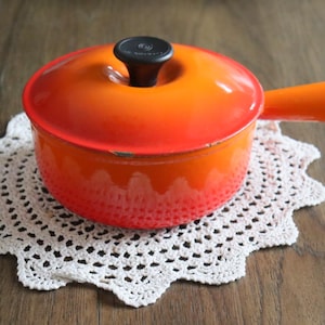May include: An orange cast iron casserole pot with a black handle and lid knob, resting on a white, crocheted doily. The pot has a single orange handle and a matching lid. The pot is approximately 15 cm in diameter.