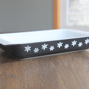 May include: Rectangular black and white baking dish with a snowflake pattern. The dish has a white interior and a black exterior with white snowflake designs. The dish is on a wooden surface.