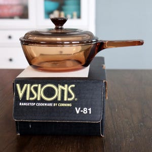 May include: A Visions amber-colored saucepan with a lid and handle, resting on a black box. The box reads "VISIONS" and "RANGETOP COOKWARE BY CORNING" with the model number V-81.