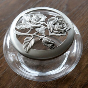 May include: A clear glass jar with a decorative metal lid. The lid features a floral design with two roses, a bud, and leaves. The jar is round and sits on a wooden surface.
