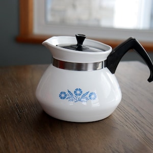 May include: White ceramic teapot with a black handle and lid. The teapot has a silver band and a blue floral design. The teapot is on a wooden surface.