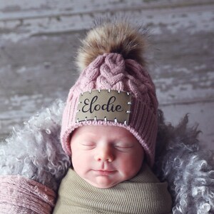 Peut inclure: Un nouveau-n&eacute; envelopp&eacute; dans une couverture kaki, portant un bonnet en tricot rose avec un pompon en fausse fourrure et une &eacute;tiquette en bois avec le nom "Elodie". Le b&eacute;b&eacute; est nich&eacute; dans un panier en osier blanc doubl&eacute; de fausse fourrure grise et de tissu rose.