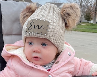 May include: A baby wearing a beige cable knit hat with two brown pom-poms and the name "Evie" on a patch. The baby is in a stroller and wearing a pink jacket and hot pink pants.