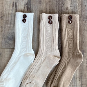 Peut inclure: Trois paires de chaussettes texturées, crème, beige et marron. Chaque chaussette présente un détail de bouton décoratif au niveau du poignet. Les chaussettes sont disposées sur une surface en bois.