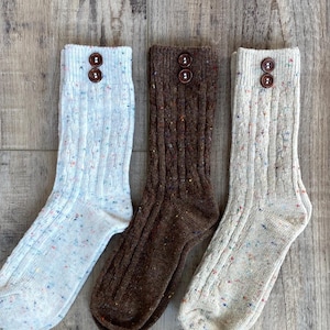 Peut inclure: Trois paires de chaussettes en tricot côtelé bleu clair, marron et beige. Chaque chaussette a un détail de bouton en haut. Les chaussettes ont un motif moucheté et sont présentées sur une surface en bois.