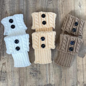 May include: Knit leg warmers in white, cream, and brown, each with a cable knit cuff and two black buttons. The leg warmers are arranged on a wooden surface, showcasing their texture and design.