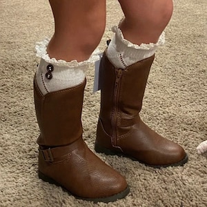 Peut inclure: Bottes en cuir marron avec fermeture éclair latérale et boucle. Les bottes sont portées avec des chaussettes blanches garnies de dentelle et de boutons décoratifs. Les bottes sont sur une surface moquettée.
