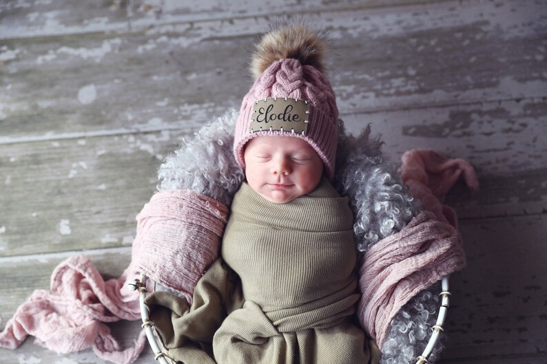Peut inclure: Un nouveau-n&eacute; envelopp&eacute; dans une couverture kaki, portant un bonnet en tricot rose avec un pompon en fausse fourrure et une &eacute;tiquette en bois avec le nom "Elodie". Le b&eacute;b&eacute; est nich&eacute; dans un panier en osier blanc doubl&eacute; de fausse fourrure grise et de tissu rose.