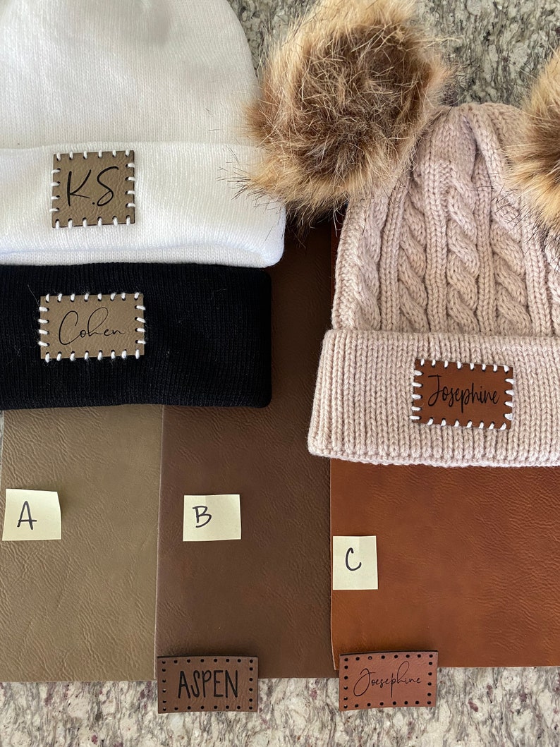 Peut inclure: Trois bonnets en blanc, noir et beige avec des &eacute;cussons en cuir personnalis&eacute;s. Le bonnet blanc porte l'inscription "KS". Le bonnet noir porte l'inscription "Cohen". Le bonnet beige porte l'inscription "Josephine".
