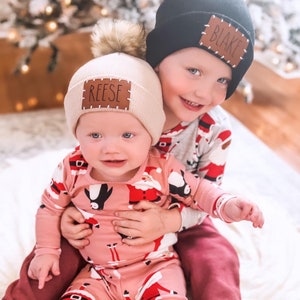 Peut inclure: Deux enfants portant des bonnets d'hiver avec des pompons et des &eacute;cussons nominatifs personnalis&eacute;s. L'un porte un bonnet noir avec "BLAKE" sur l'&eacute;cusson, l'autre un bonnet beige avec "REESE". Les deux enfants portent des pyjamas sur le th&egrave;me de No&euml;l. L'enfant avec le bonnet beige est tenu par l'autre enfant.