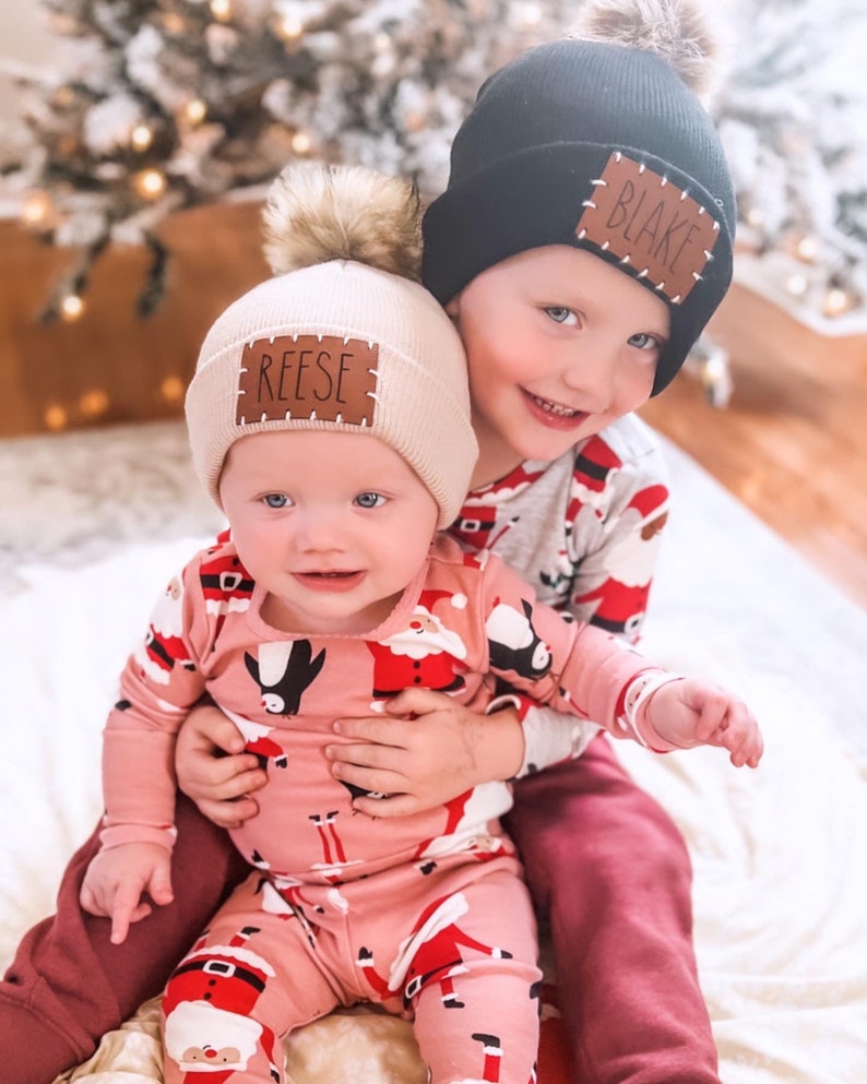 Peut inclure: Deux enfants portant des bonnets d'hiver avec des pompons et des &eacute;cussons nominatifs personnalis&eacute;s. L'un porte un bonnet noir avec "BLAKE" sur l'&eacute;cusson, l'autre un bonnet beige avec "REESE". Les deux enfants portent des pyjamas sur le th&egrave;me de No&euml;l. L'enfant avec le bonnet beige est tenu par l'autre enfant.
