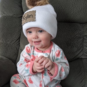 Peut inclure: Un bonnet en tricot blanc avec un pompon en fausse fourrure marron. Le bonnet porte une &eacute;tiquette en cuir marron avec le nom "Kylin" cousu dessus. Le b&eacute;b&eacute; porte une combinaison &agrave; motifs floraux.