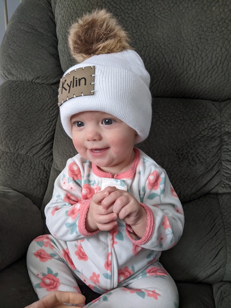 Peut inclure: Un bonnet en tricot blanc avec un pompon en fausse fourrure marron. Le bonnet porte une &eacute;tiquette en cuir marron avec le nom "Kylin" cousu dessus. Le b&eacute;b&eacute; porte une combinaison &agrave; motifs floraux.