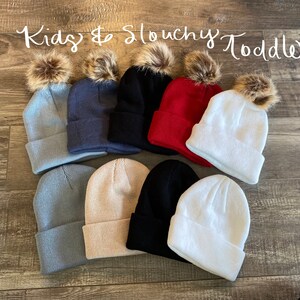 Peut inclure: Plusieurs bonnets slouchy pour enfants et tout-petits, de diff&eacute;rentes couleurs : bleu clair, marine, noir, rouge, blanc et beige. Chaque bonnet est dot&eacute; d'un bord repli&eacute; et d'un pompon en fausse fourrure marron. Le texte "Kids & Slouchy Toddler" est &eacute;crit en cursive.