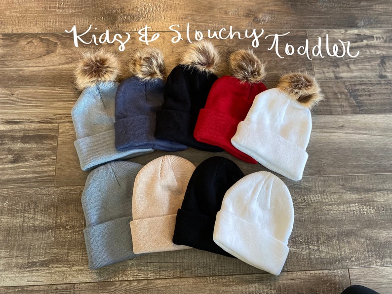 Peut inclure: Plusieurs bonnets slouchy pour enfants et tout-petits, de diff&eacute;rentes couleurs : bleu clair, marine, noir, rouge, blanc et beige. Chaque bonnet est dot&eacute; d'un bord repli&eacute; et d'un pompon en fausse fourrure marron. Le texte "Kids & Slouchy Toddler" est &eacute;crit en cursive.