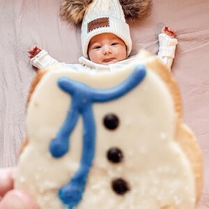 Peut inclure: Un b&eacute;b&eacute; portant un bonnet blanc en tricot torsad&eacute; avec deux pompons en fausse fourrure marron et le nom "Addison" sur un patch marron. Un biscuit en forme de bonhomme de neige avec une &eacute;charpe bleue et des boutons noirs est au premier plan.