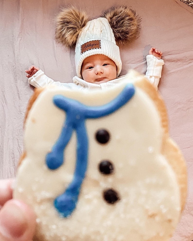 Peut inclure: Un b&eacute;b&eacute; portant un bonnet blanc en tricot torsad&eacute; avec deux pompons en fausse fourrure marron et le nom "Addison" sur un patch marron. Un biscuit en forme de bonhomme de neige avec une &eacute;charpe bleue et des boutons noirs est au premier plan.