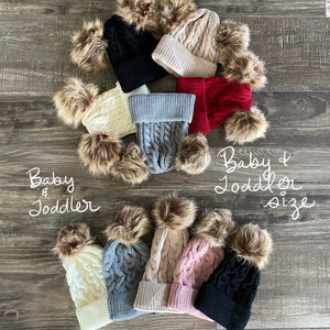 Peut inclure: Plusieurs bonnets pour b&eacute;b&eacute;s et tout-petits en tricot torsad&eacute;, de couleurs vari&eacute;es : noir, cr&egrave;me, gris, beige, rouge et rose. Chaque bonnet est orn&eacute; d'un pompon en fausse fourrure. Le texte "Baby & Toddler size" est visible.