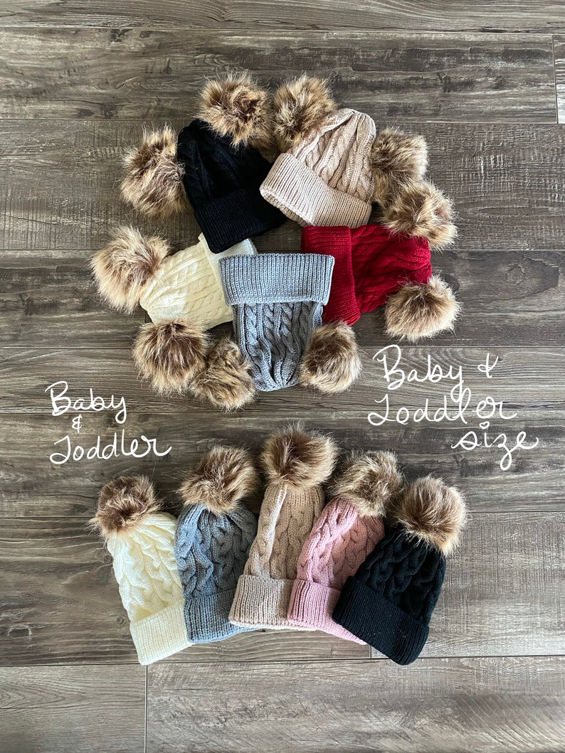 Peut inclure: Plusieurs bonnets pour b&eacute;b&eacute;s et tout-petits en tricot torsad&eacute;, de couleurs vari&eacute;es : noir, cr&egrave;me, gris, beige, rouge et rose. Chaque bonnet est orn&eacute; d'un pompon en fausse fourrure. Le texte "Baby & Toddler size" est visible.